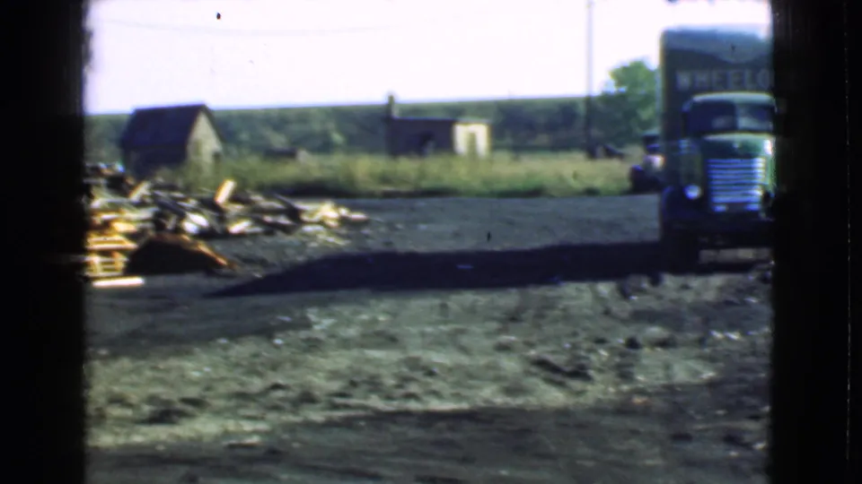 Cleveland, Ohio home-movie frame showing the green truck near the side of the yard, with scrap piles, utility poles, and small rural buildings.