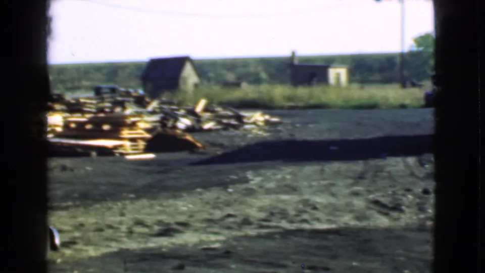 1939 Cleveland-area home-movie frame showing a partial maroon sedan near lumber stacks, sheds, dirt ground, and a distant tree line.