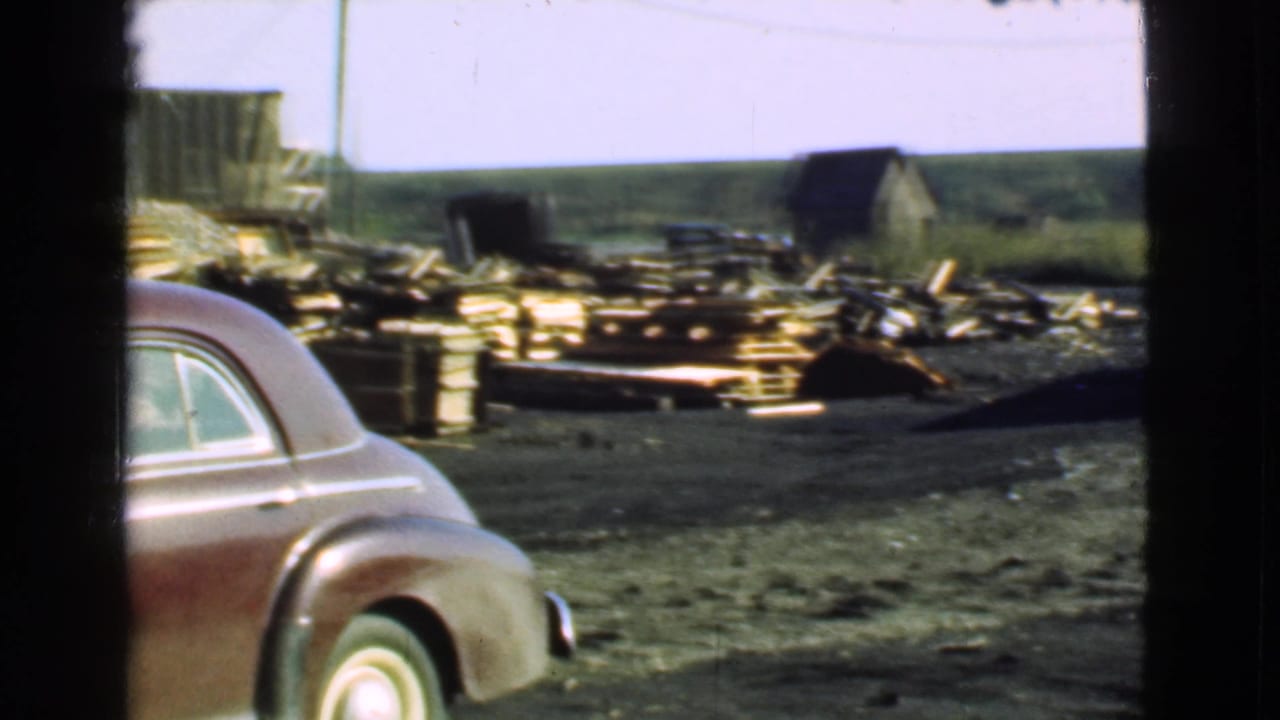 Maroon sedan parked in front of lumber piles and yard buildings.