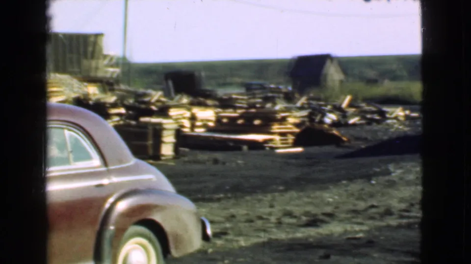 1939 Cleveland-area frame with the rear side of a maroon sedan in front of lumber stacks, open ground, small sheds, and utility poles.
