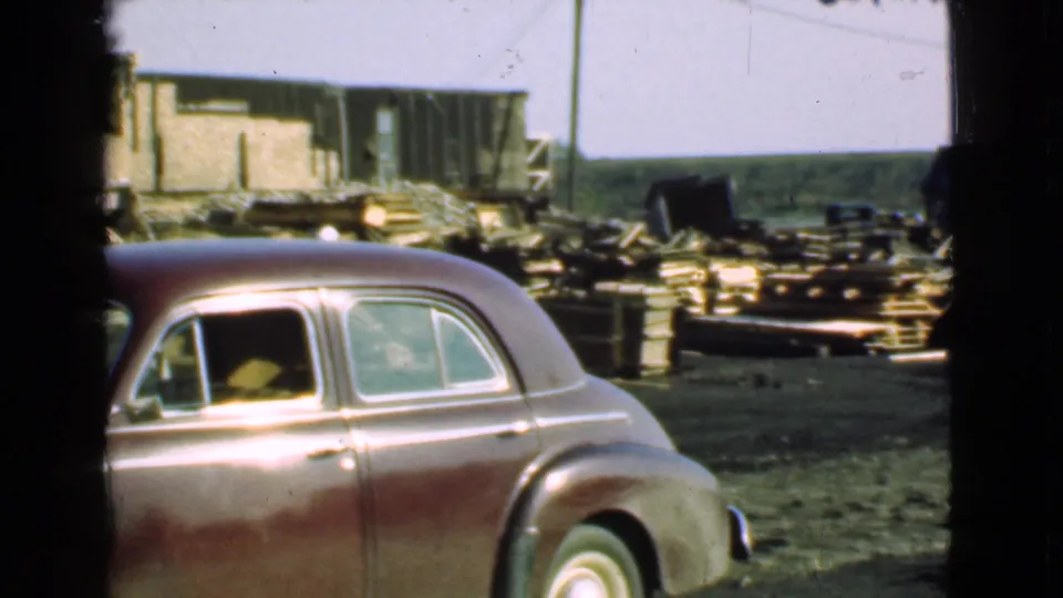 1939 Cleveland-area home-movie frame showing the side of a maroon sedan against a lumber yard with stacked boards, sheds, and open ground.
