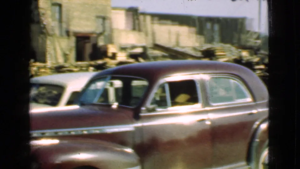 1939 Cleveland-area frame showing a maroon sedan beside a pale car, with stacked lumber, low yard buildings, and open ground behind them.