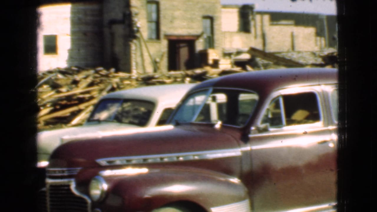 1939 Cleveland home-movie frame showing a maroon sedan beside a pale car near a weathered house and stacked demolition debris.