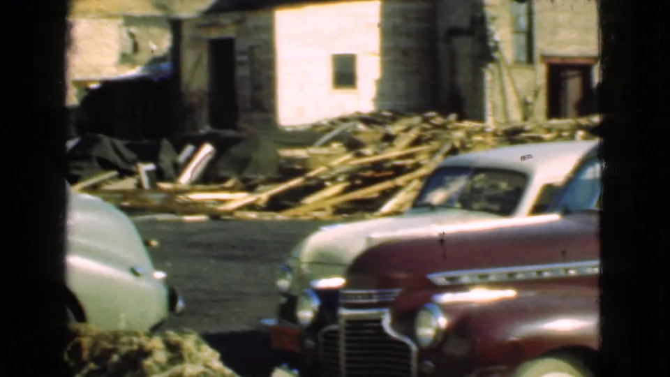 1939 Cleveland frame with a pale coupe and maroon sedan partially visible against a weathered masonry house and stacked wood debris.