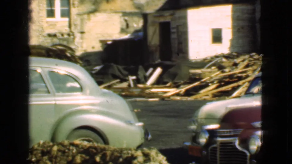 1939 Cleveland home-movie view of a pale green coupe and maroon sedan near rubble, lumber, and a weathered residential structure.