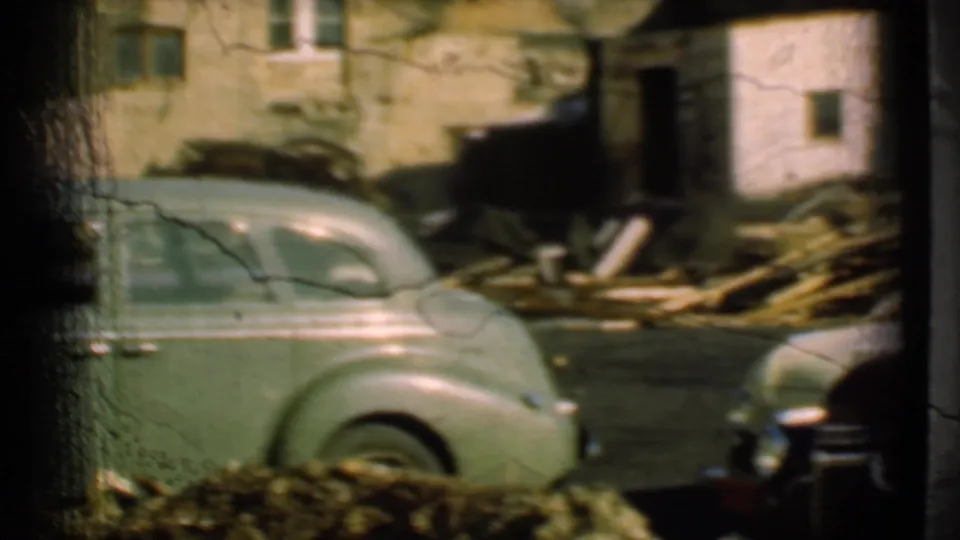 Soft 8mm frame from 1939 Cleveland showing a pale green coupe, part of a maroon car, dark framing edges, and debris near an old house.