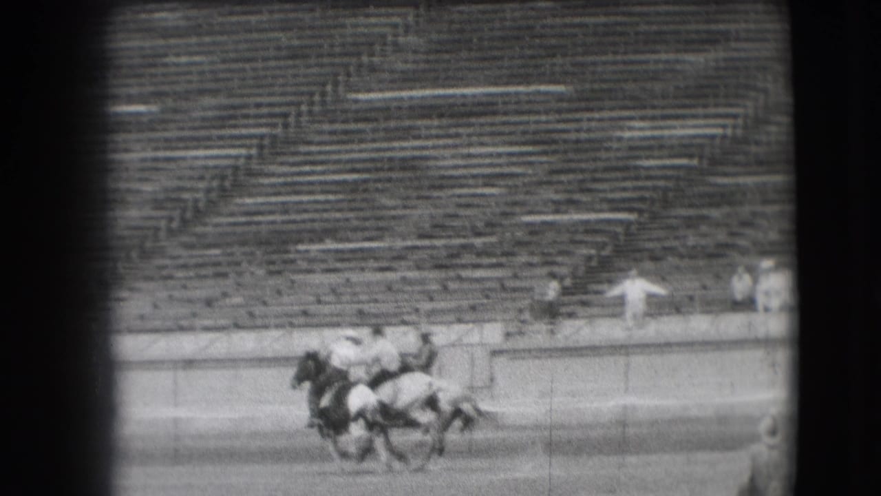 Cowboys ride horses across the arena in front of empty bleachers.