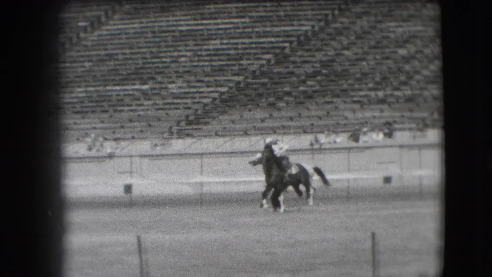 A pair of riders on horseback crosses the Denver arena while mostly empty stadium bleachers and the arena wall remain visible behind them.