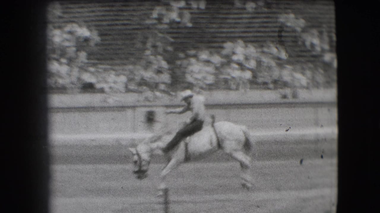 Cowboy rides a light-colored bucking horse in a rodeo arena.