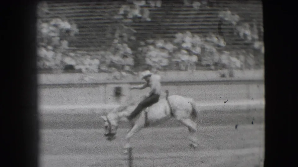 In 1941 Denver rodeo footage, a cowboy leans back while a light-colored horse bucks near the arena rail.