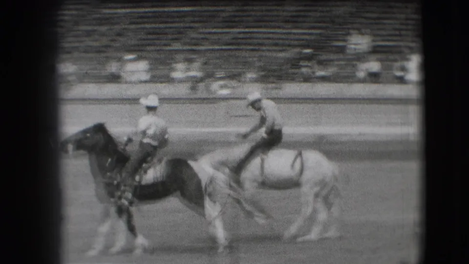 Two riders on horseback are seen close together in the 1941 Denver rodeo arena, with stands behind them.