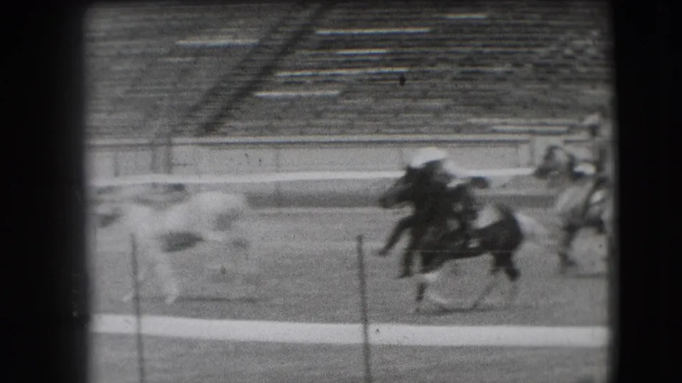 1941 Denver horse-track frame with horses running past a low figure near the rail, filmed with blur and heavy grain.