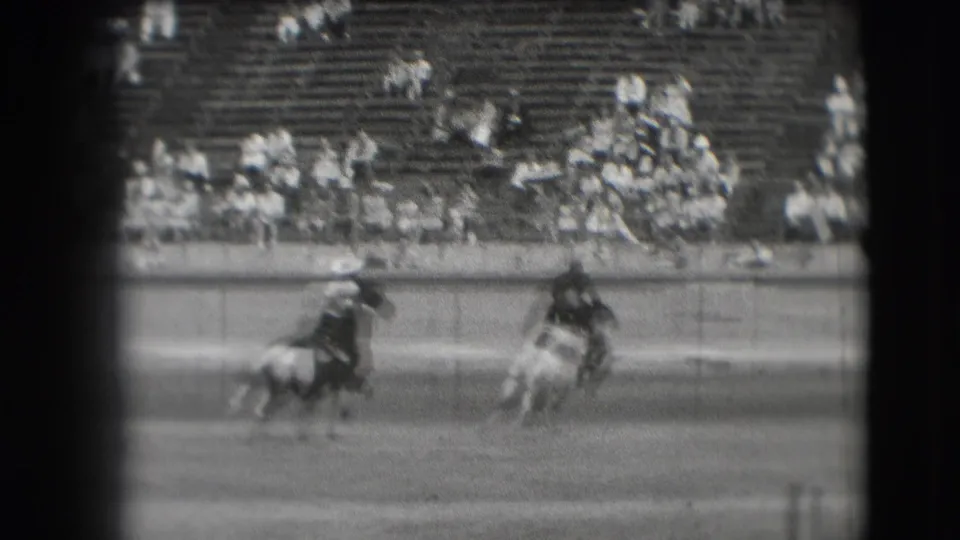 Two rodeo riders move through the Denver stadium arena, one low on a bucking horse as spectators watch from the bleachers.