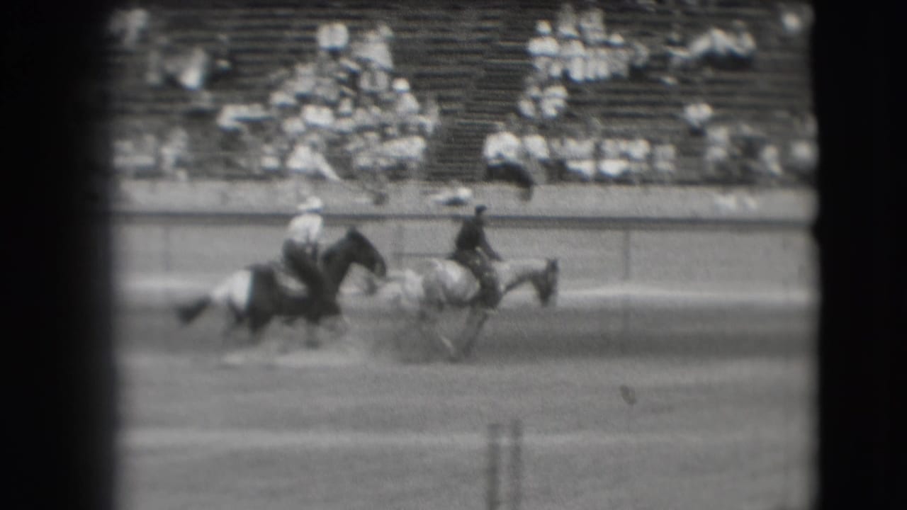 Two riders on horseback move across a dirt arena with spectators seated in bleachers behind them.