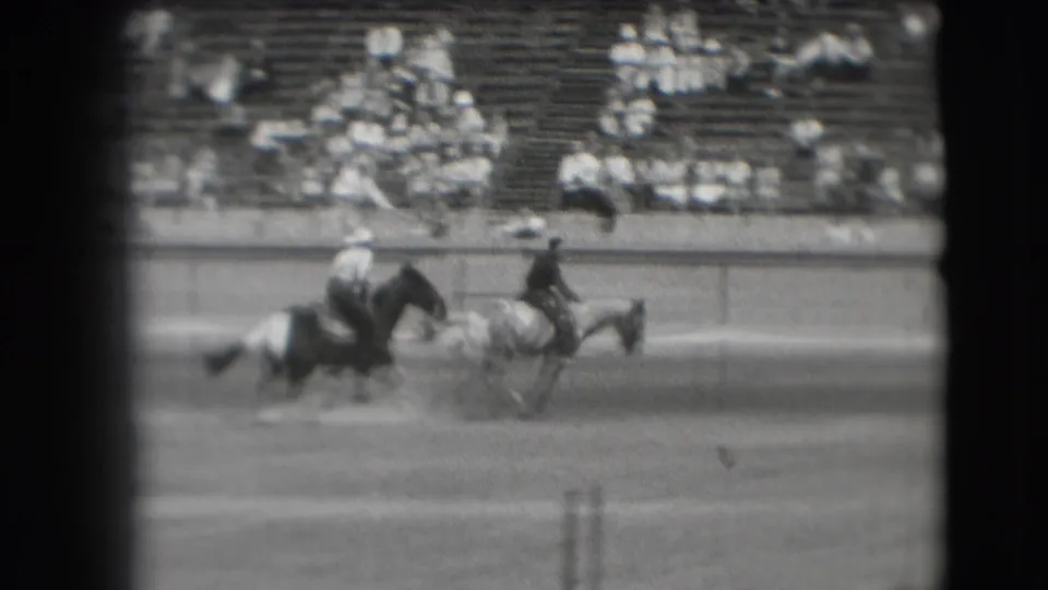 1941 Denver rodeo home-movie frame showing horseback riders crossing a dirt arena while distant spectators watch from bleachers.