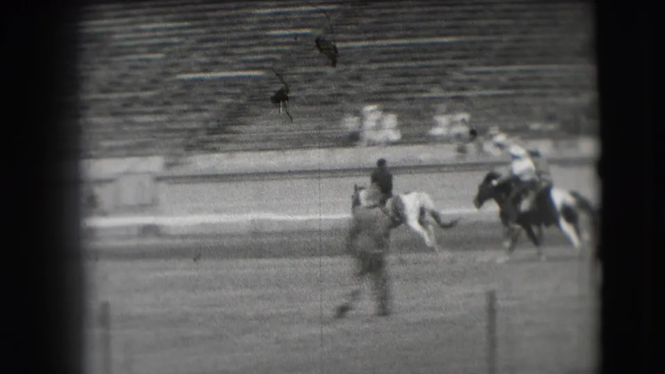 Two mounted riders travel across the arena with a foreground figure blurred by motion and spectator seating behind them.