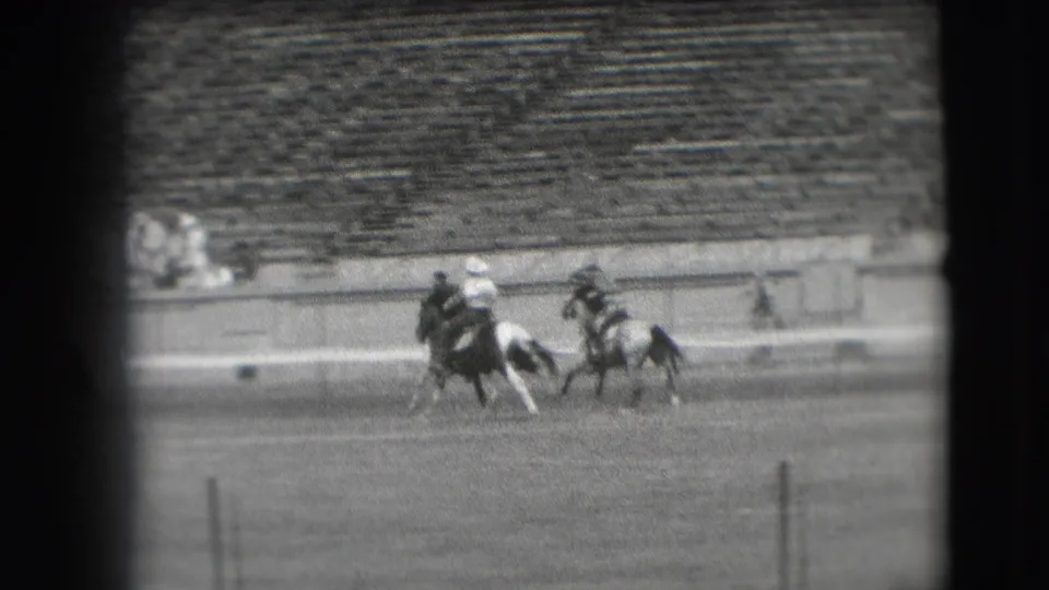Two mounted riders gallop across the arena in black-and-white home-movie footage, with tiered bleachers visible behind them.