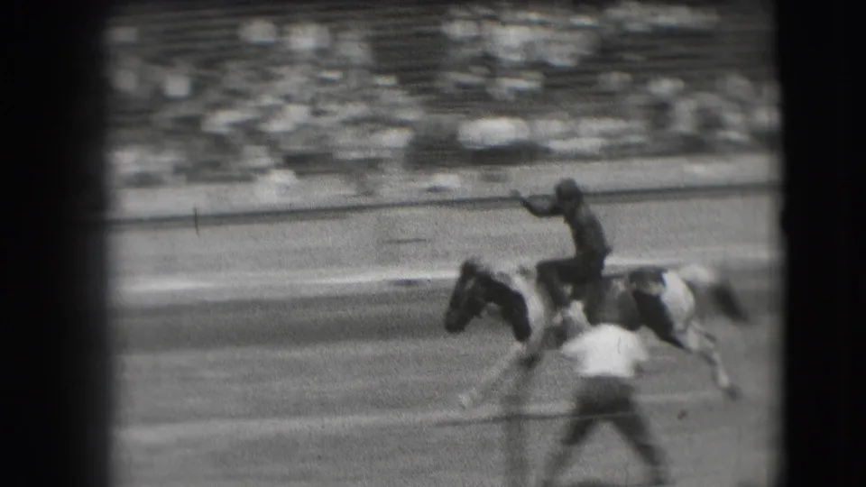The rider continues on the bucking pinto as a second arena figure appears in the foreground of the spectator-shot frame.
