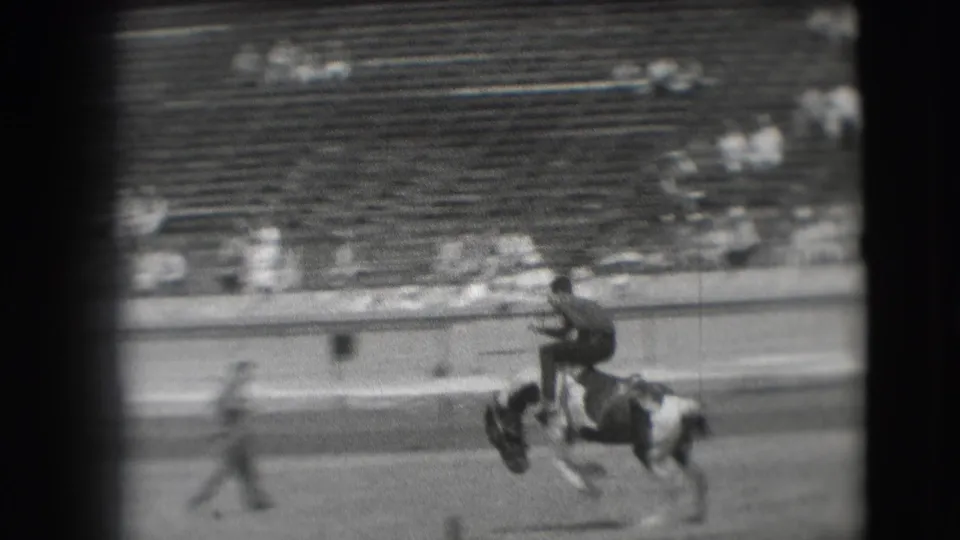 In 1941 Denver footage, a rider leans forward on a bucking pinto horse while distant stadium seats fill the background.