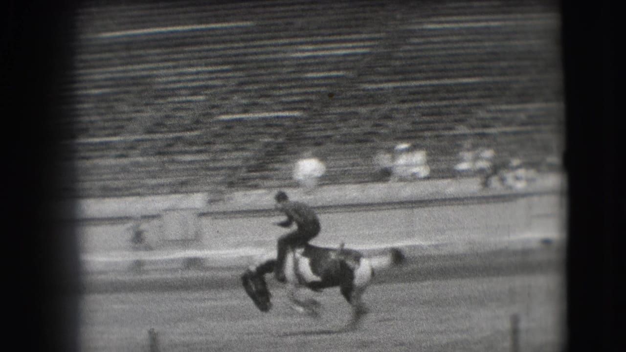 A cowboy appears to ride a bucking pinto horse across the Denver stadium arena, seen from a distant spectator angle.