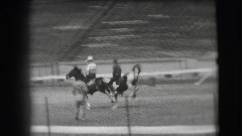 A rider in a light hat moves beside another mounted figure in the Denver stadium arena, with seating visible behind them.