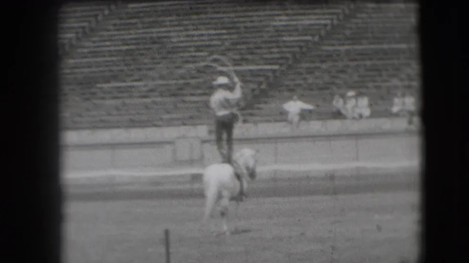 A cowboy in the 1941 Denver arena raises an arm while balancing on a white horse before the bleachers.
