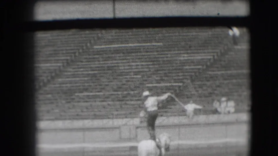 In 1941 Denver, a cowboy in a light hat stands on a white horse and swings a lasso before mostly empty bleachers.