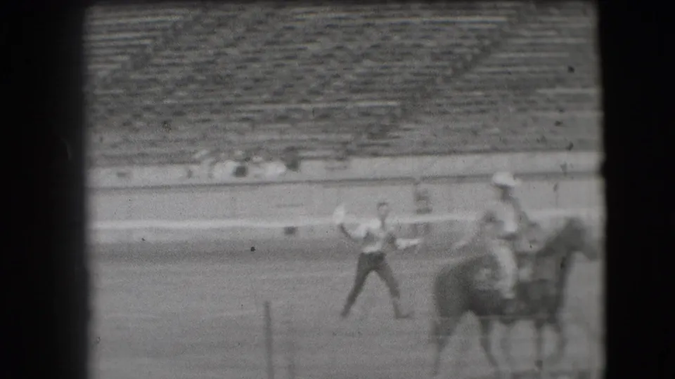 A cowboy holds the rope near horses at the side of the arena, with bleachers and a few distant spectators behind them.