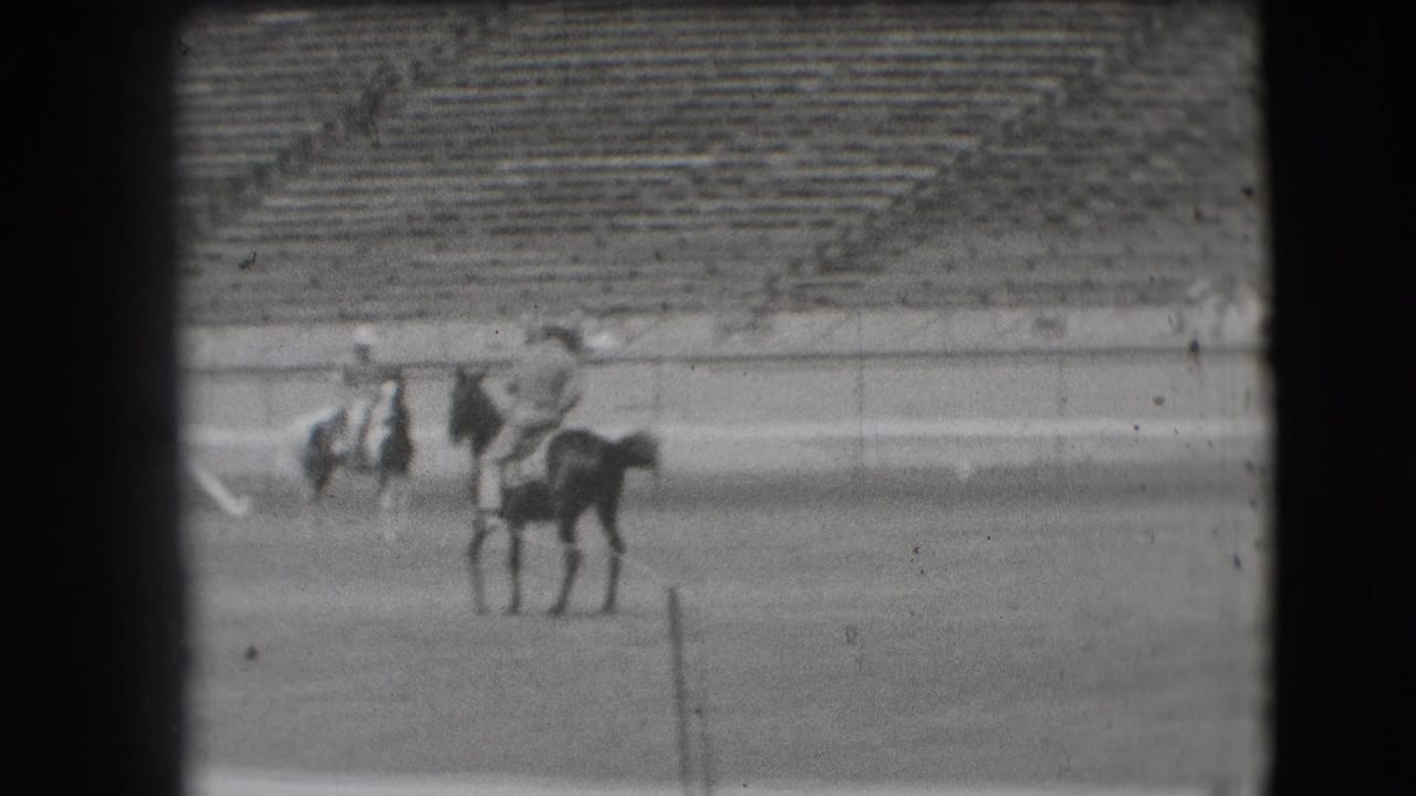 1941 Denver rodeo home-movie frame showing riders on horses in an outdoor arena with empty and occupied stadium seats behind.