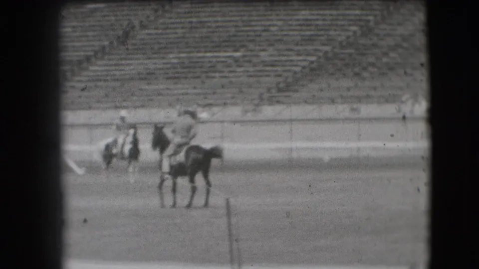 1941 Denver rodeo home-movie frame showing riders on horses in an outdoor arena with empty and occupied stadium seats behind.