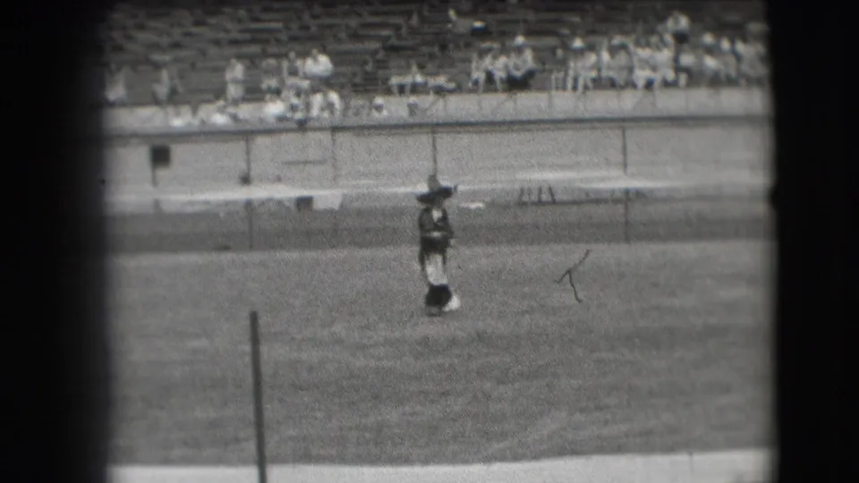 1941 Denver rodeo home-movie still showing a cowboy in hat and chaps standing alone in the arena with a rope visible nearby.