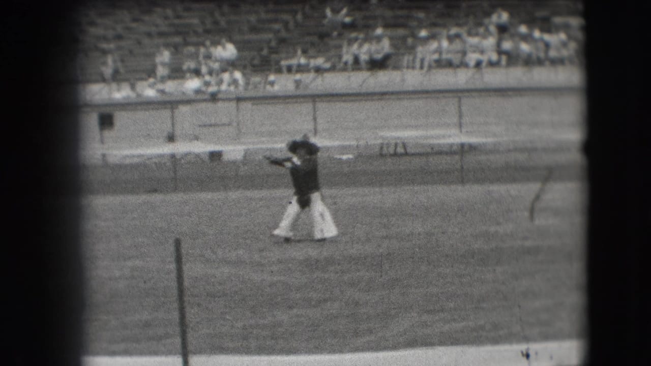 Cowboy-style dancer crossing a grassy stadium field with spectators in the bleachers.