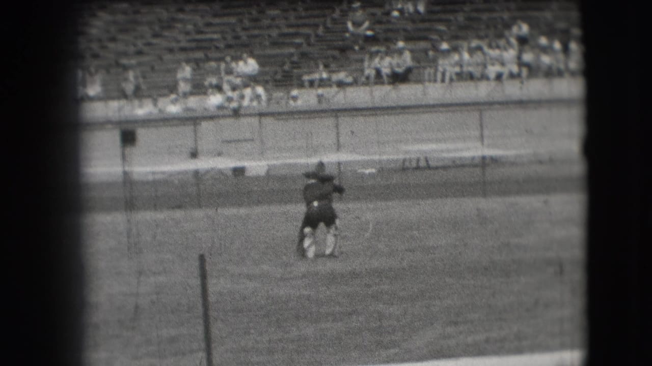 Costumed dancer bends or pivots on the field near a stadium barrier and bleachers.