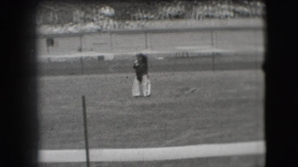 Black-and-white 8mm frame from Denver showing the clown paused on the grass below the stands.
