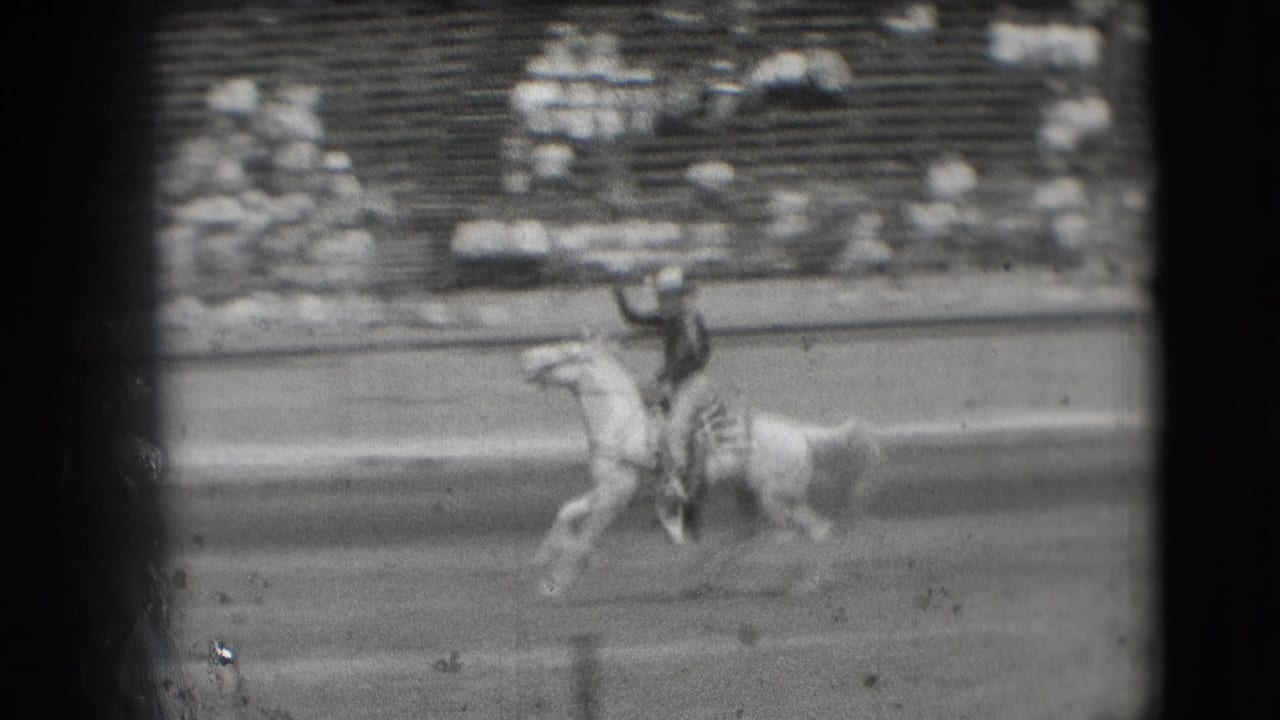 Cowboy on a light-colored horse gallops across a rodeo arena with spectators in the stands.