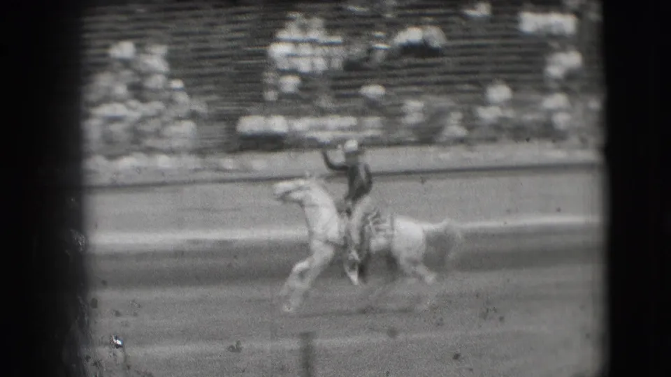 In 1941 Denver rodeo footage, a cowboy rides a light-colored horse across the arena while raising one arm near tiered seating.