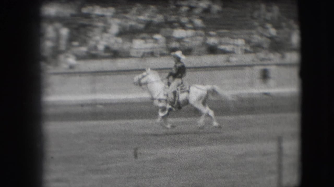 The rider continues across the Denver arena on a light-colored horse, with blurred spectators visible in the background seating.