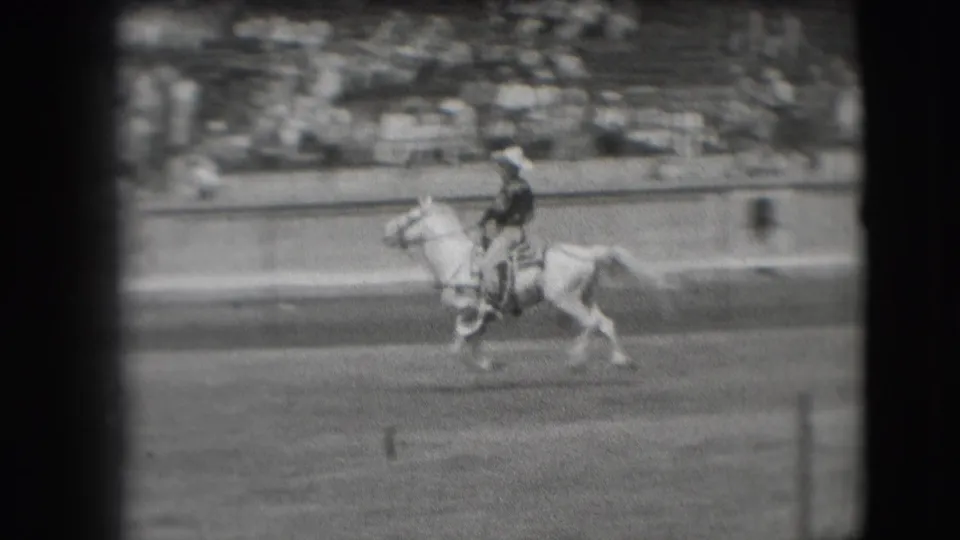 The rider continues across the Denver arena on a light-colored horse, with blurred spectators visible in the background seating.