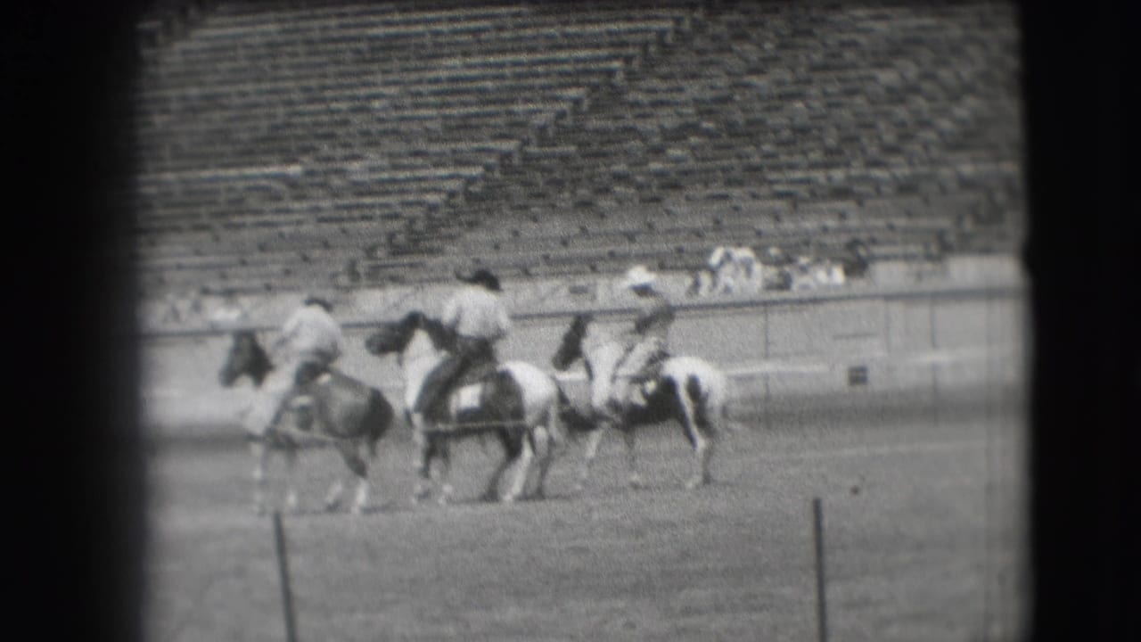 1941 Denver rodeo home-movie frame with three riders on horses crossing the field before rows of spectators.