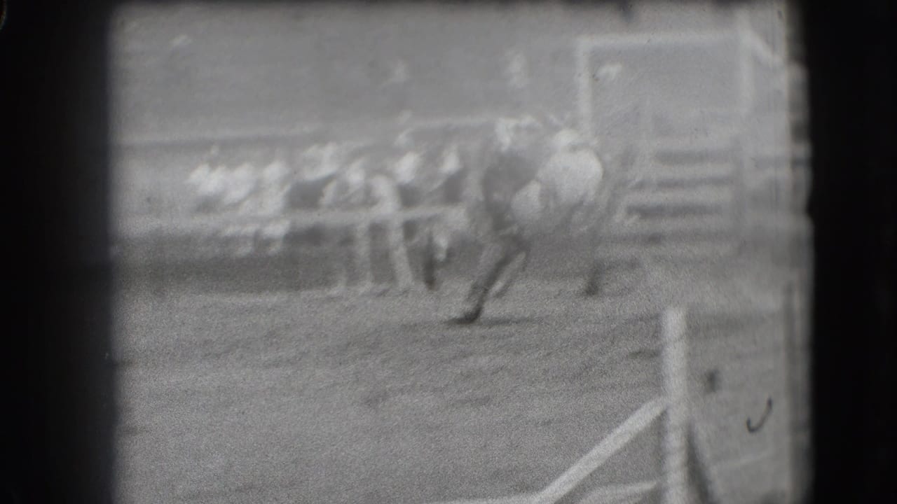 In Denver archival 8mm footage, a rider appears on a bucking horse inside a rodeo arena with spectators beyond the rail.