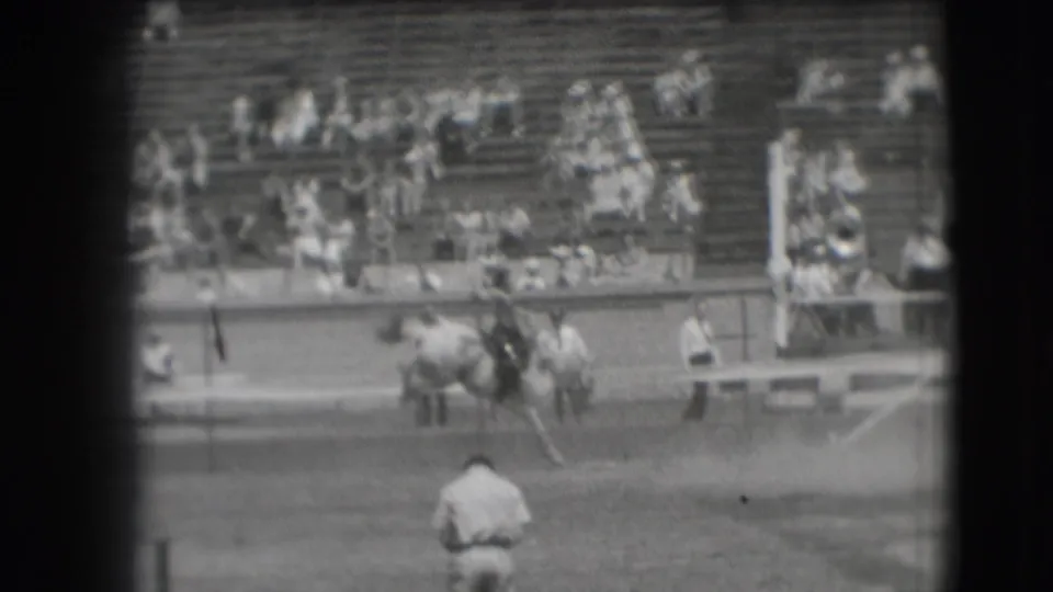 The rider and bucking horse turn near the Denver rodeo arena rail, with a seated crowd visible across the bleachers.