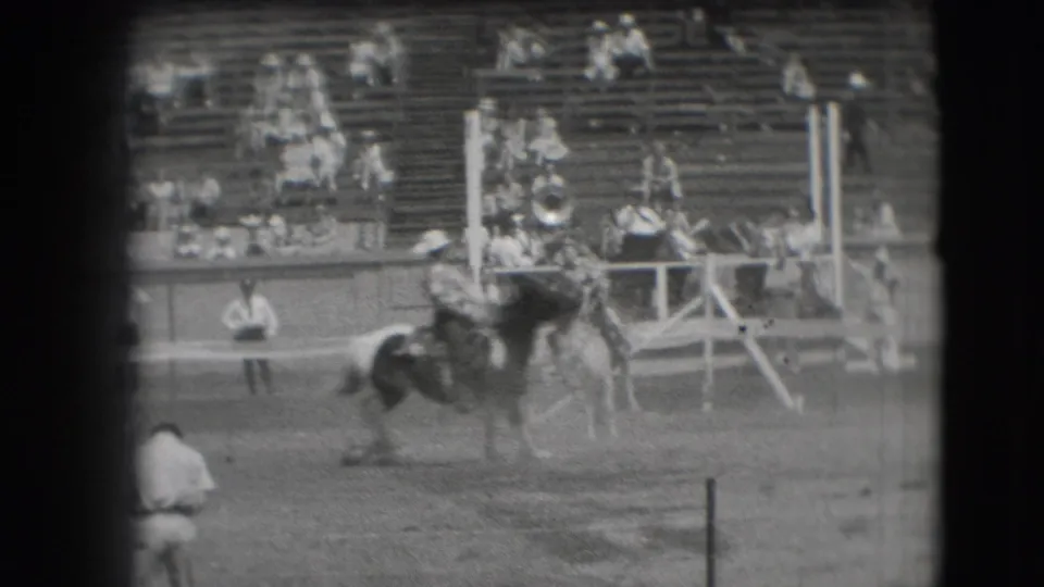 Black-and-white 8mm frame of a rodeo horse and rider near the rail, with Denver grandstand spectators in the background.