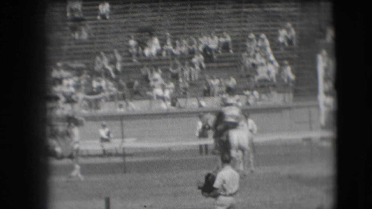 Soft black-and-white 8mm frame of a Denver rodeo rider passing the arena rail as spectators sit in the grandstand.