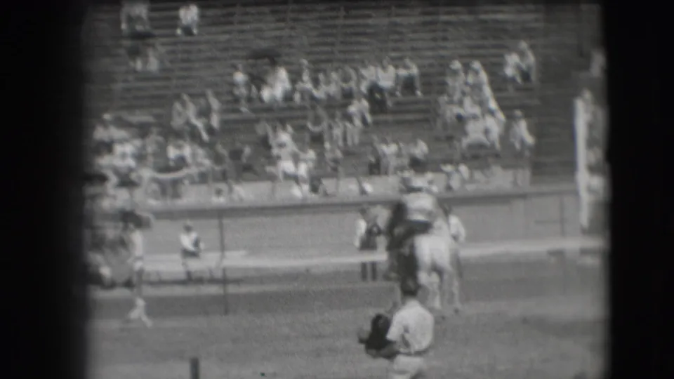 Soft black-and-white 8mm frame of a Denver rodeo rider passing the arena rail as spectators sit in the grandstand.