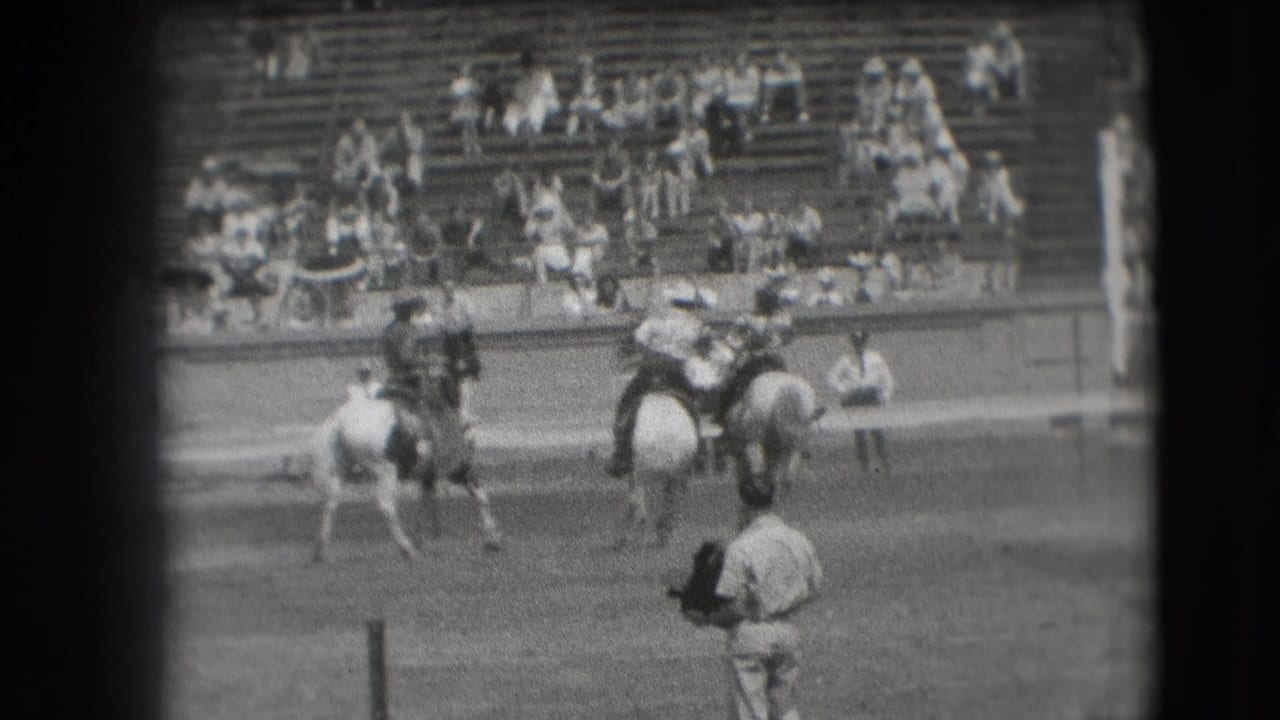 Rodeo riders on horseback move across an arena with spectators in bleachers behind them.