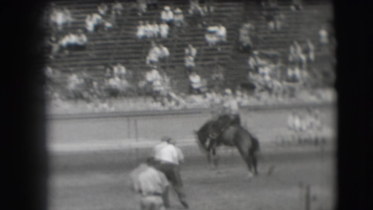 Bucking horse with rider in a rodeo arena as handlers and spectators watch.