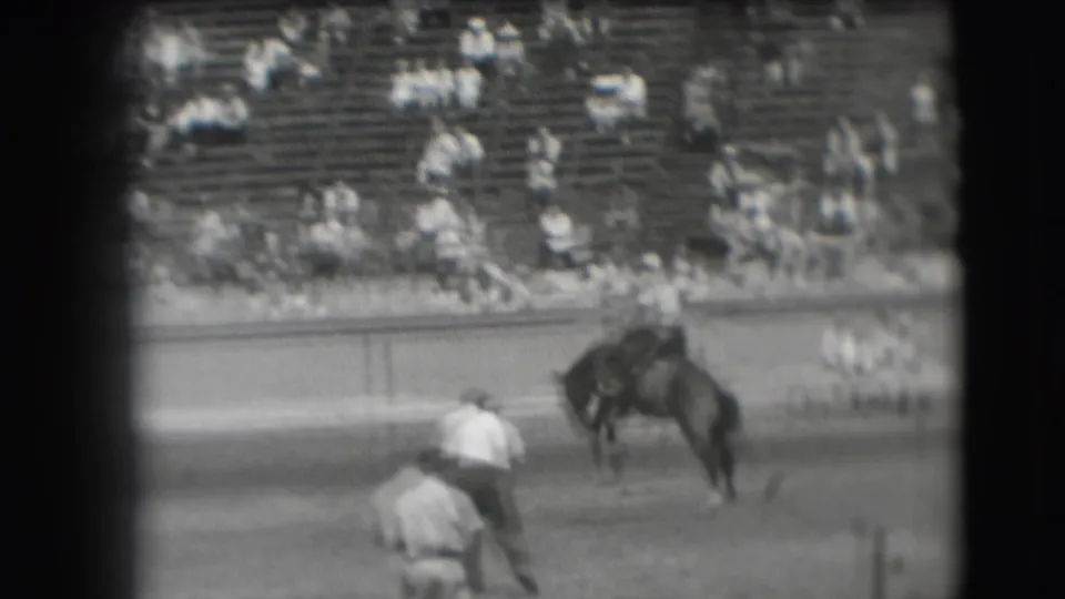 In 1941 Denver rodeo footage, a rider leans over a bucking horse while arena handlers and bleacher spectators remain in the background.