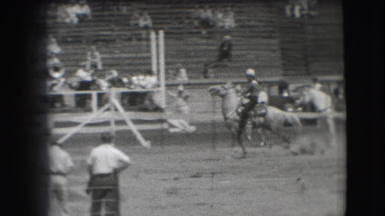 A rider holds on as a horse bucks near the rail in the Denver rodeo arena, with officials or handlers and spectators visible behind.