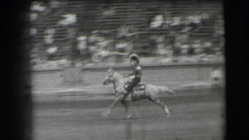 The rodeo rider continues across the dirt arena in the 1941 Denver home movie, with blurred bleachers and spectators behind the action.