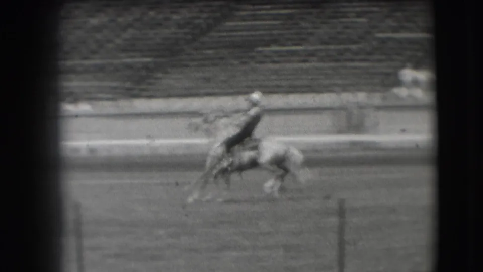 A mounted competitor rides across the Denver rodeo arena, with distant bleachers and spectators in the background.